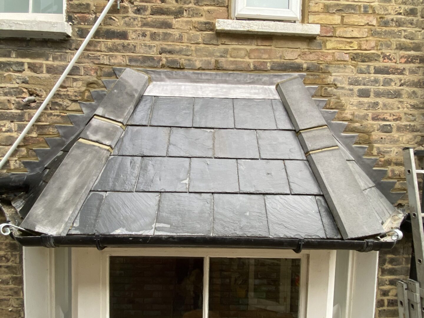 a slate roof awning above what appears to be the entrance to a building. The slates are arranged in an overlapping pattern and are predominantly grey in color. Metal flashings line the edges of the awning, and there is a brick wall visible in the background