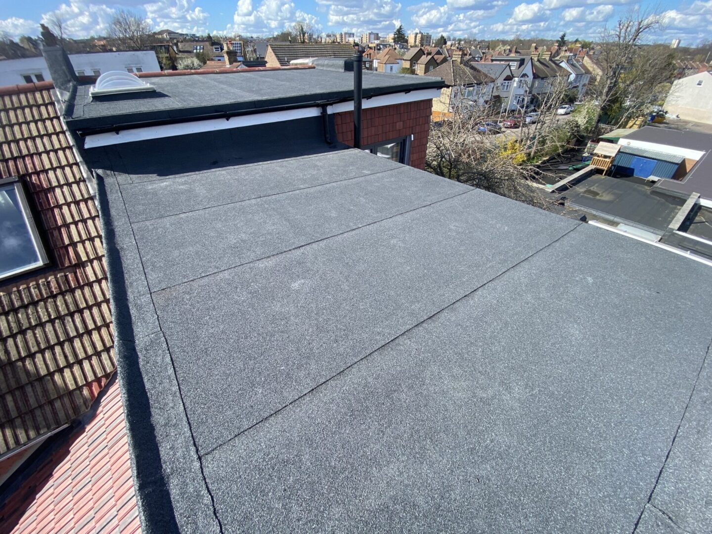 A rooftop view with textured surface, vent pipe, and suburban houses under a clear blue sky.