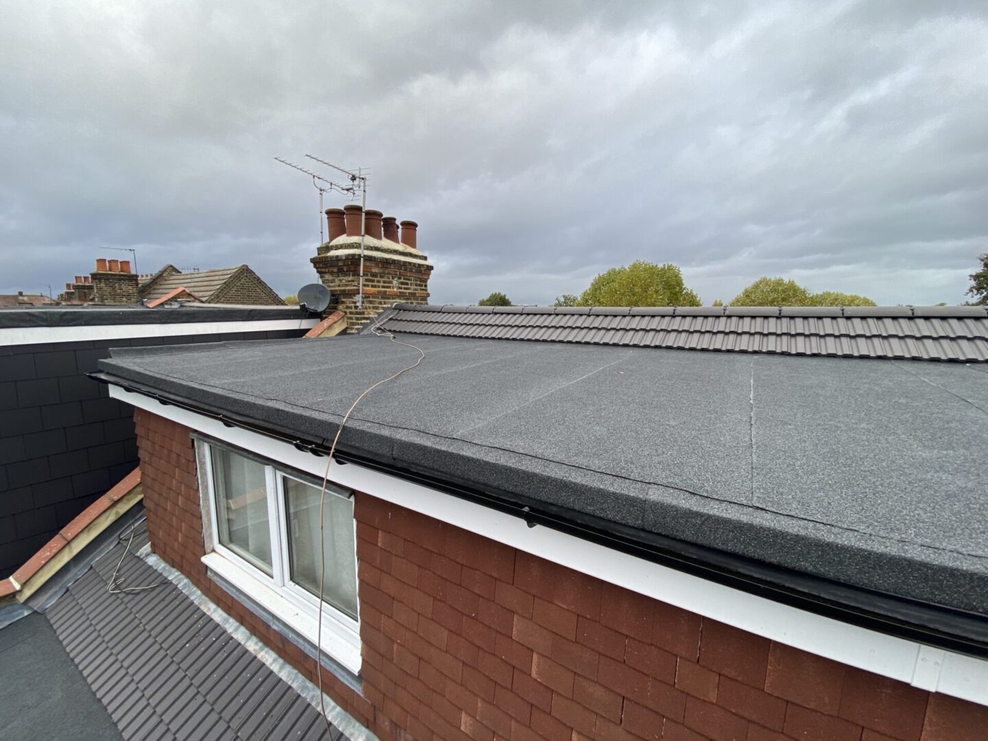 A rooftop view featuring a flat roof with a black covering, leading to a brick chimney stack against a cloudy sky. Below is a window of a building with white frames and red brick walls.