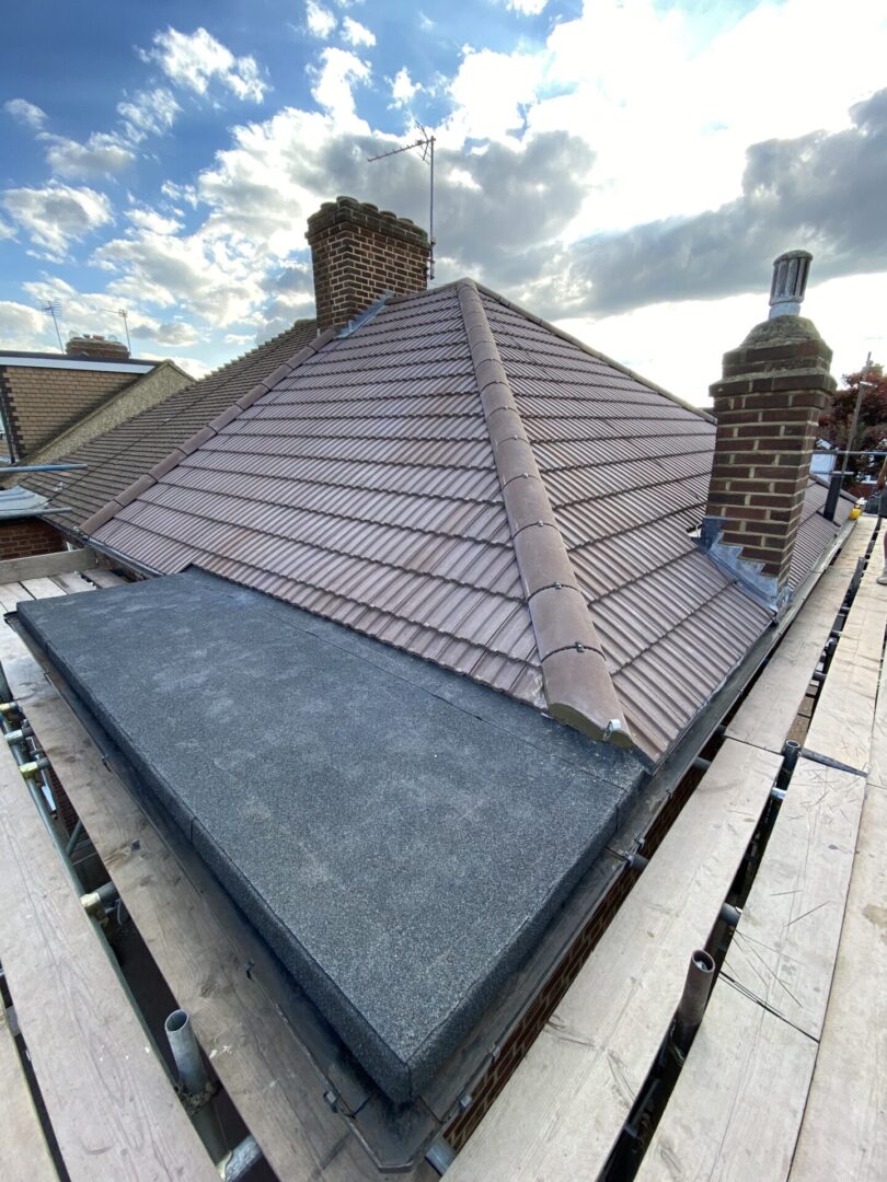 A close-up view of a grey tiled roof with multiple slopes and ridges, featuring a chimney on the left side, under a blue sky with scattered clouds. The roof is bordered by scaffolding at the bottom edge, indicating construction or repair work.