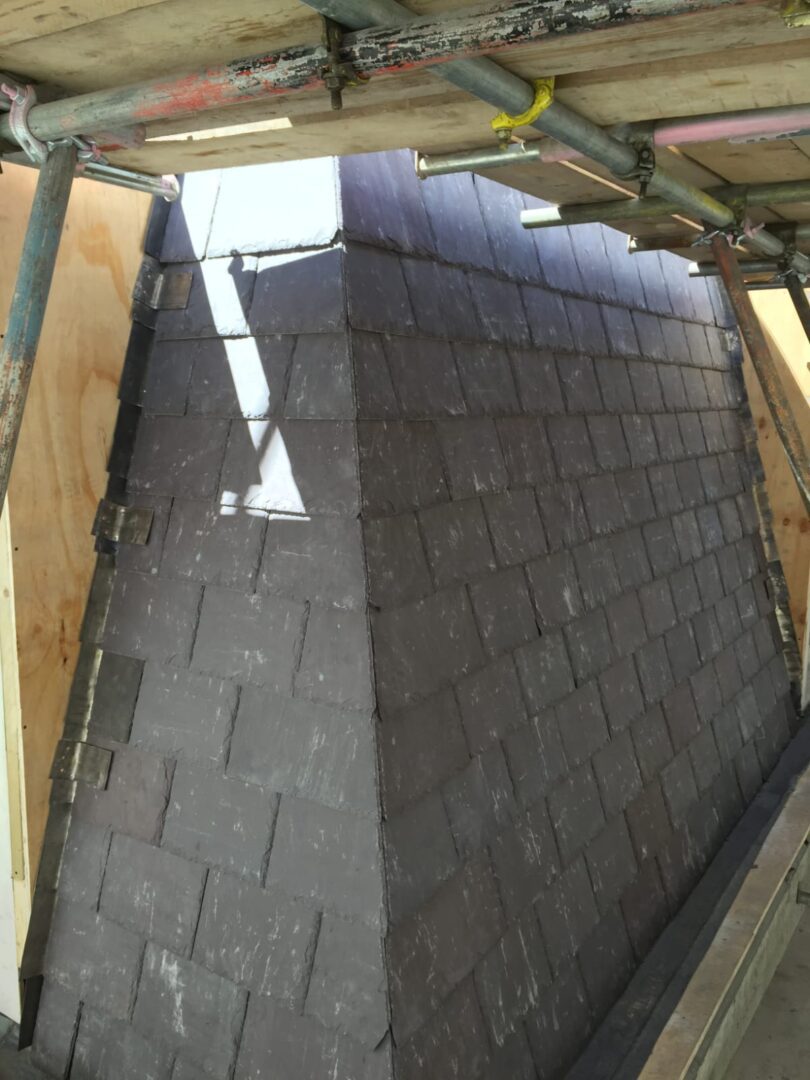 A rooftop under construction with dark shingles partially installed, wooden scaffolding visible.