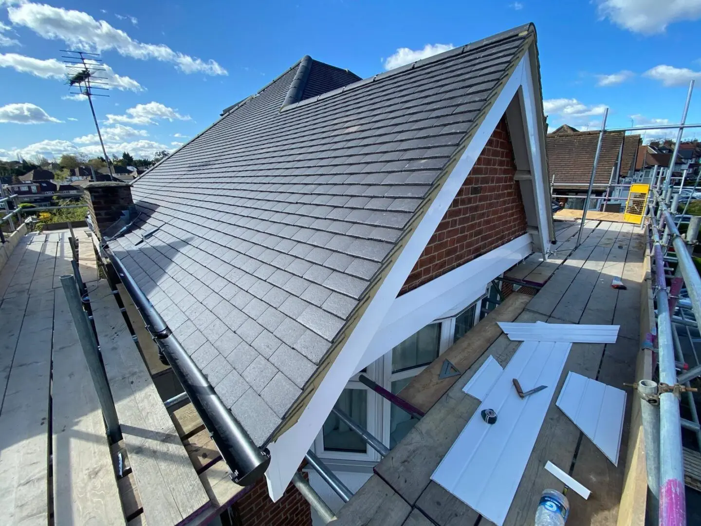 A newly constructed steep-sloped roof with grey shingles and black gutters, surrounded by scaffolding, under a bright blue sky with scattered clouds.