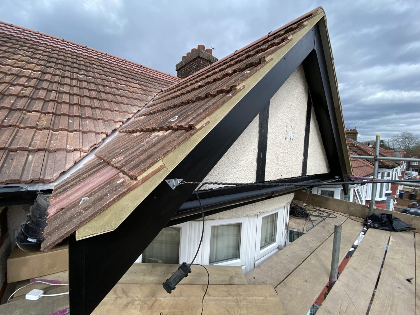 a house under construction with exposed roof beams, surrounded by other houses with red tiled roofs under an overcast sky.